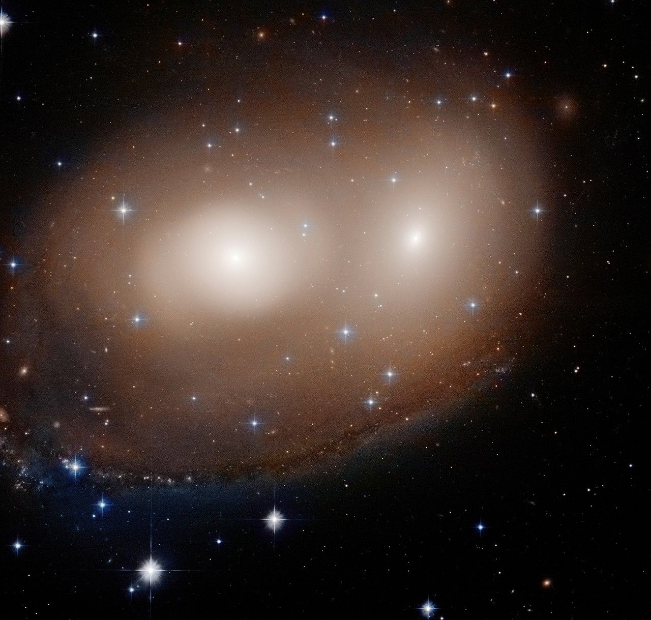 Two galaxies with bright white centers and white glows around them look like eyes surrounded by a reddish haze of gas and dust, with a brownish glow of the same forming an arc around them, thicker on the bottom.